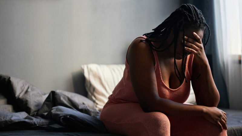 Mulher negra sentada na cama com as mãos sobre o rosto, demonstrando sofrimento emocional